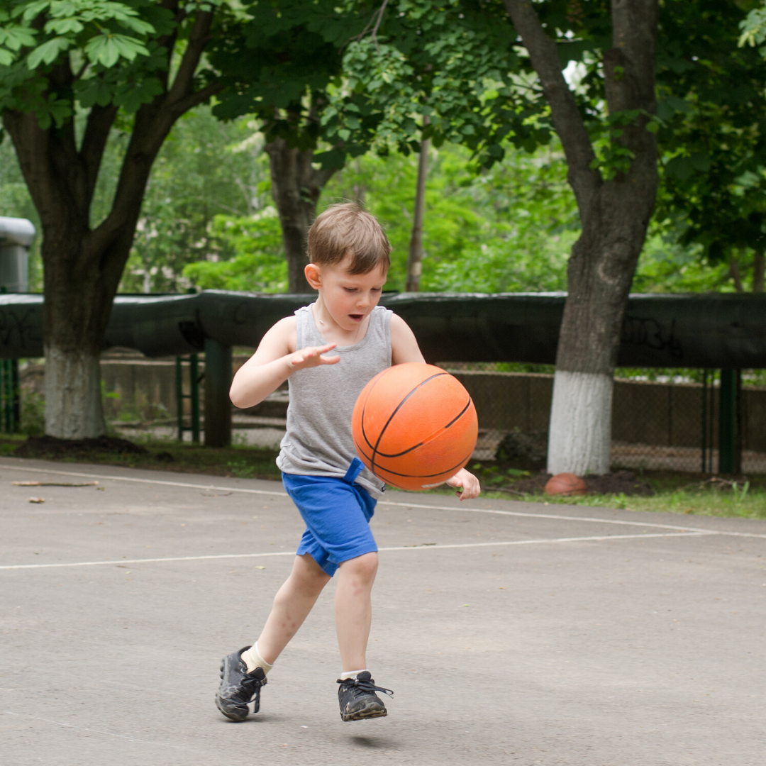 Little boy dribbling basketball on pavement surrounded by trees